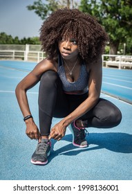 Young Black Woman Athlete With Afro Hair Looking To The Side Crouching Tying Her Shoelaces With Black Leggings, Gray Top, Gray And Pink Sneakers On Blue Running Track And Background Trees