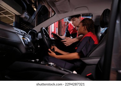 Young Black woman automotive mechanic technician and partner checking maintenance list with tablet in car interior at garage. Vehicle service fix and repair works, industrial occupation business jobs.
