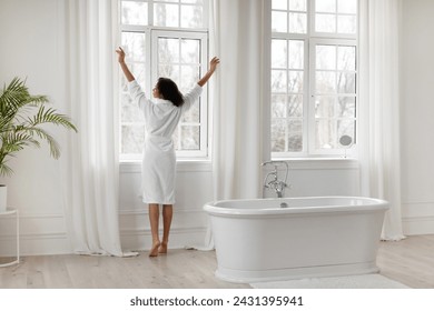 Young black woman in bathrobe opening curtains in light bathroom, lady standing near big window, ready for beauty treatments, enjoy morning routine - Powered by Shutterstock