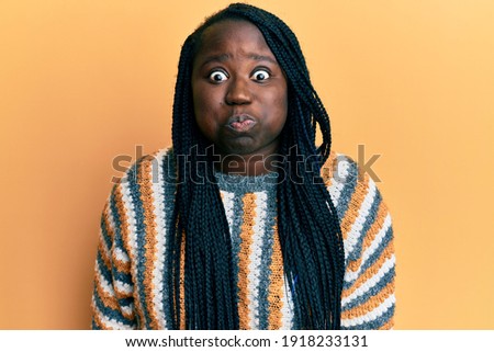 Young black woman with braids wearing casual winter sweater puffing cheeks with funny face. mouth inflated with air, crazy expression. 