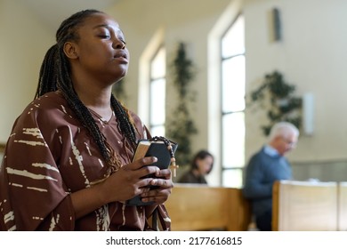 Young Black Woman In Casual Dress Keeping Her Eyes Closed During Silent Prayer In Church While Holding Holy Bible And Rosary Beads