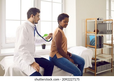 Young Black Woman Comes To Hospital For Health Checkup. Male Doctor At Modern Clinic Sitting On Medical Exam Couch With Female Patient And Using Stethoscope To Check Her Breathing Or Heartbeat