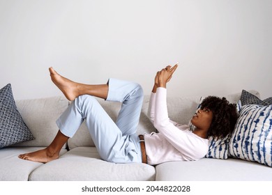 Young Black Woman With Curly Hair Laying Down On Couch Using Her Phone