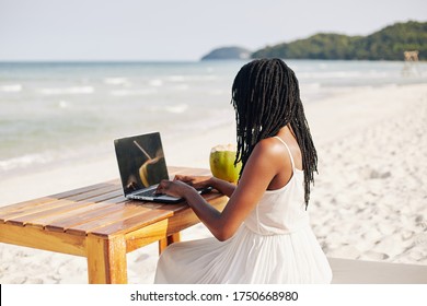 Young Black Woman With Dreadlocks Sitting At Wooden Table On Beach And Working On Laptop