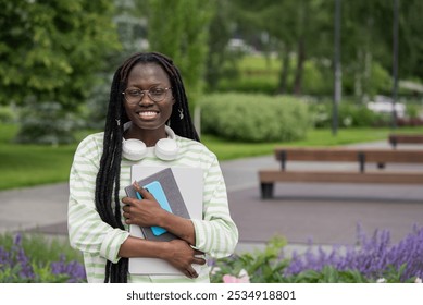 Young black woman with dreadlocks is smiling at camera while holding laptop, smartphone and headphones. She is enjoying her time during break between classes - Powered by Shutterstock