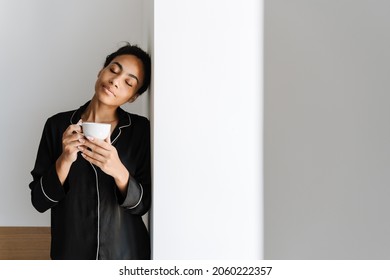 Young Black Woman Drinking Tea While Leaning On Wall At Home