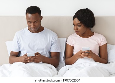 Young Black Woman Eavesdropping On Her Boyfriend Using Mobile Phone, Looking At His Smartphone Screen. African American Couple Sitting In Bed And Using Mobile Phones. Distrust In Relationships