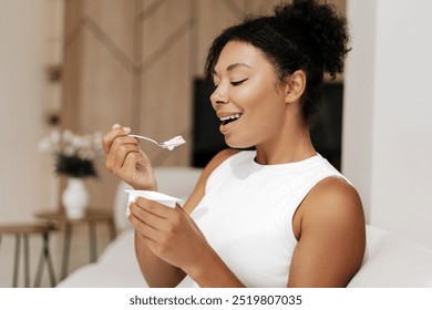 Young black woman enjoys yogurt on her couch at home, embodying healthy eating habits with a cheerful smile - Powered by Shutterstock