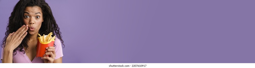 Young Black Woman Expressing Surprise While Eating French Fries Isolated Over Purple Background