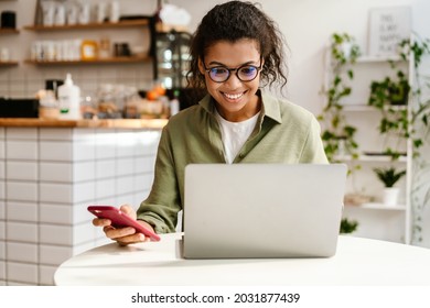 Young Black Woman In Eyeglasses Using Cellphone While Working With Laptop At Cafe Indoors