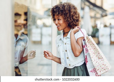 Young black woman in front of a shop window in a shopping street. African girl with afro hairstyle wearing casual clothes. - Powered by Shutterstock