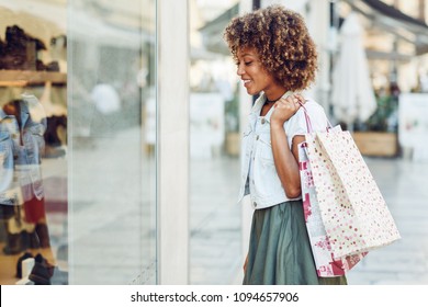 Young Black Woman In Front Of A Shop Window In A Shopping Street. African Girl With Afro Hairstyle Wearing Casual Clothes.