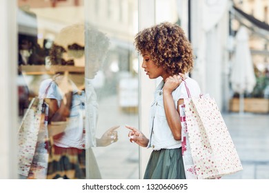 Young Black Woman In Front Of A Shop Window In A Shopping Street. African Girl With Afro Hairstyle Wearing Casual Clothes.