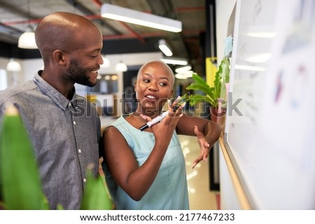 A young Black woman gestures to the whiteboard while planning with colleague