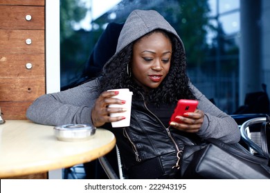 Young Black Woman in Gray Jacket with Hood Sitting on Chair Having Coffee While Busy Texting.