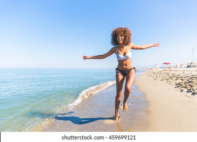 Young Black Woman Having Fun At Seaside. She Is Twenty Years Old, Mixed Race Caucasian And African Black, With Curly And Voluminous Hair, Running With Open Arms And Happy Face.