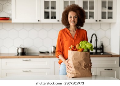 Young Black Woman In Kitchen Posing With Grocery Bag After Food Shopping, Happy African American Female Holding Package With Fresh Vegetables And Fruits, Enjoying Healthy Eating, Copy Space - Powered by Shutterstock