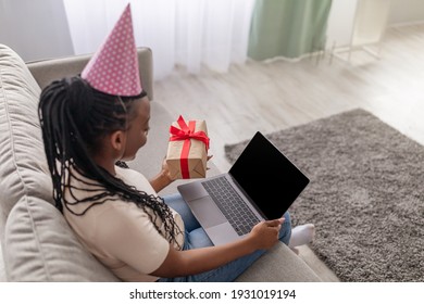 Young Black Woman With Party Hat And Present Using Laptop With Empty Screen At Home, Copy Space. African American Lady Having Online Party, Showing Gift Box To Blank Computer Screen, Mockup