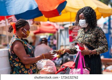 Young black woman paying money to a vendor for a purchased item in the market. Two women wearing locally made mask and surgical mask on the street in covid-19 pandemic season for protection.