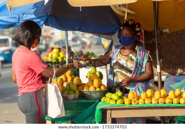 Young black woman paying money to a fruit vendor\
for a purchased item in the market. Two women wearing locally made\
masks on the street in covid-19 pandemic season for protection -\
Marketing in Africa