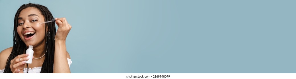 Young Black Woman With Pigtails Smiling While Applying Face Serum Isolated Over Blue Background