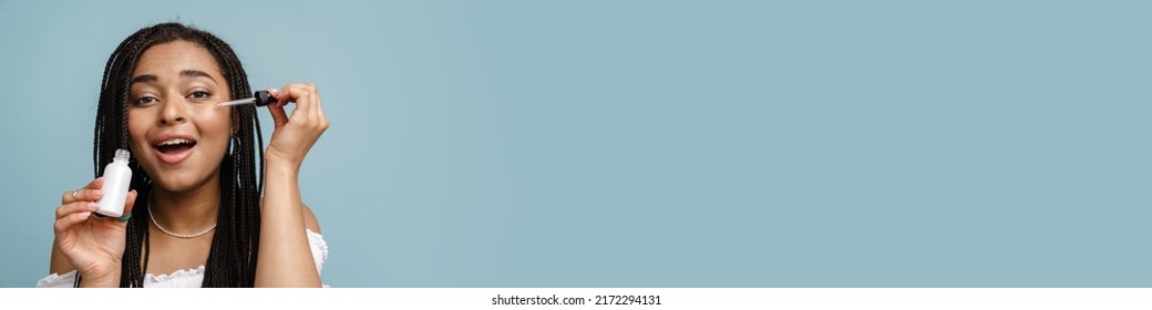 Young Black Woman With Pigtails Smiling While Applying Face Serum Isolated Over Blue Background