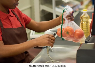 Young Black Woman In Red Shirt And Brown Apron Scanning Fresh Tomatoes In Cellophane Bag Over Cashier Counter In Supermarket