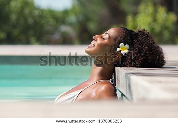 Young black woman relaxing at spa pool. Beautiful\
woman relaxing in outdoor spa swimming pool with head leaning at\
poolside. Closeup face of attractive girl with closed eyes enjoy\
vacation at resort.