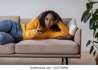 Young Black Woman With Remote Control Lying On Sofa, Feeling Bored Watching TV, Switching Channels, Tired Of Dull Movie At Home During Covid Outbreak. Domestic Cinema Concept