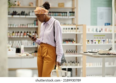 Young Black Woman With Shopping Basket Scrolling In Smartphone While Standing In Front Of Display With Cosmetic Products In Supermarket