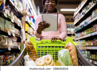 Young Black Woman With Shopping Cart Full Of Products Looking Into Her Grocery List At Supermarket. Lovely African American Lady Buying Products At Mall, Low Angle View. Selective Focus