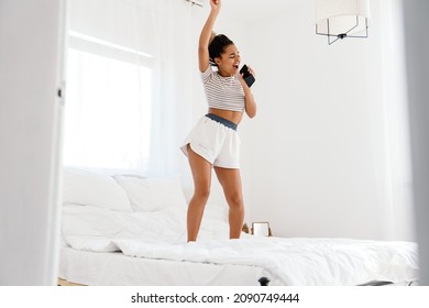 Young Black Woman Singing While Dancing With Cellphone On Bed At Home