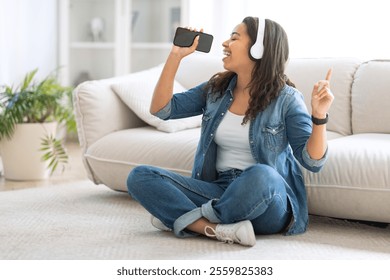 A young black woman sits on the floor with crossed legs, wearing headphones and singing along to music from her smartphone. She appears happy and engaged in her personal moment. - Powered by Shutterstock