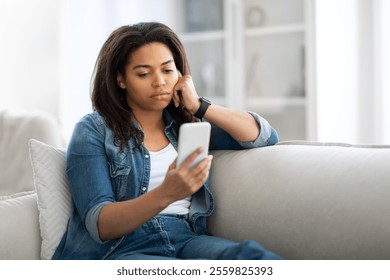 A young black woman sits on a couch in her living room, looking intently at her smartphone. She wears a denim jacket and appears deep in thought while relaxing in her cozy space. - Powered by Shutterstock