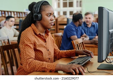 Young Black woman sitting at desk in university library working on desktop computer and listening to music in headphones - Shutterstock ID 2160229077