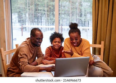 Young black woman sitting in front of laptop with her family and explaining something to friend during online communication