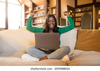 Young Black Woman Sitting On Couch With Laptop, Lifting Hands Up, Gesturing YES, Celebrating Success, Winning Online Lottery Or Casino Bet At Home. Shopping Sale, Promotion, Achievement Concept