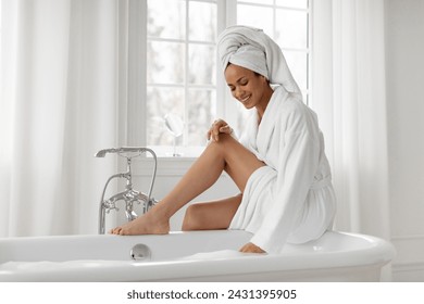 Young black woman sitting on bathtub and checking water, lady in bathrobe preparing to take bath, relaxing in bathroom at home, free space - Powered by Shutterstock