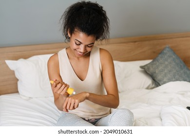 Young Black Woman Smiling And Applying Hand Cream While Sitting On Bed At Home