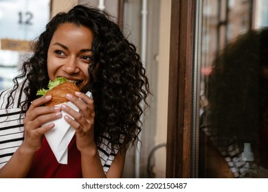 Young Black Woman Smiling And Eating Sandwich While Sitting At Cafe Outdoors