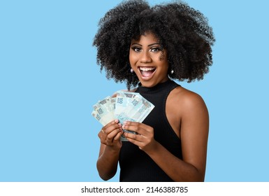 Young Black Woman Smiling Holding Brazilian Money Bills, Positively Surprised, Space For Text, Person, Advertising Concept, Isolated On Blue Background.