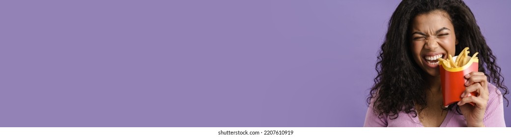 Young Black Woman Smiling While Eating French Fries Isolated Over Purple Background