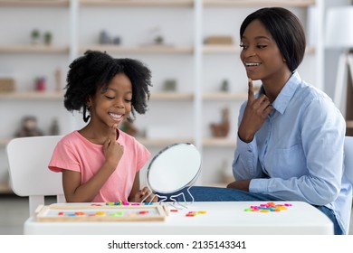 Young Black Woman Speech Pathologist Exercising With Adorable Little Girl With Language Difficulties, Working On Word Sounds, Kid Looking At Mirror, Touching Chin And Smiling, Clinic Interior