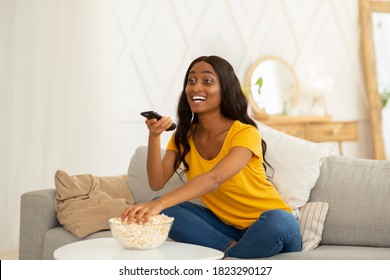 Young Black Woman Spending Her Free Time Watching TV And Eating Popcorn On Sofa At Home, Copy Space. Pretty African American Lady With Remote Control Enjoying Interesting Movie In Living Room