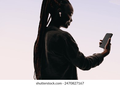 Young Black woman in a studio wears a denim jacket, styling her hair in a ponytail. Holding a smartphone, connected to earbuds, she communicates confidently online with an app for a video chat. - Powered by Shutterstock