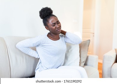 Young Black woman suffering from backache at home. Portrait of a young brunette girl sitting on the couch at home with a headache and back pain. Beautiful woman Having Spinal Or Kidney Pain - Powered by Shutterstock