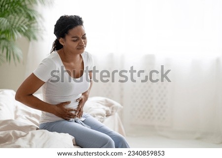 Young black woman suffering stomach pain in the morning, sitting on bed in bedroom and touching belly, african american lady in pajamas feeling unwell at home, having menstruation cramps, copy space