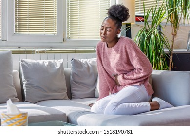 Young Black Woman Suffering From Stomachache On Sofa At Home. Woman Sitting On Bed And Having Stomach Ache. Young Woman Suffering From Abdominal Pain While Sitting On Sofa At Home