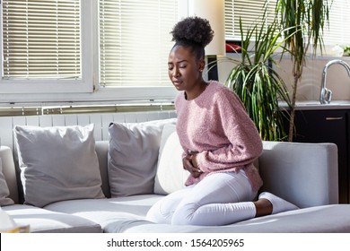 Young Black Woman Suffering From Stomachache On Sofa At Home. Woman Sitting On Bed And Having Stomach Ache. Young Woman Suffering From Abdominal Pain While Sitting On Sofa At Home