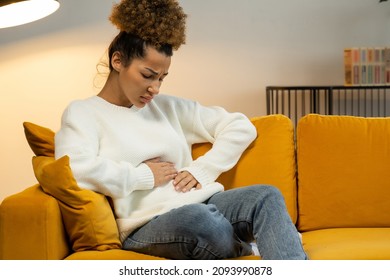 Young Black Woman Suffers From Abdominal Pain On The Couch At Home. A Woman Is Sitting On The Bed And Her Stomach Hurts